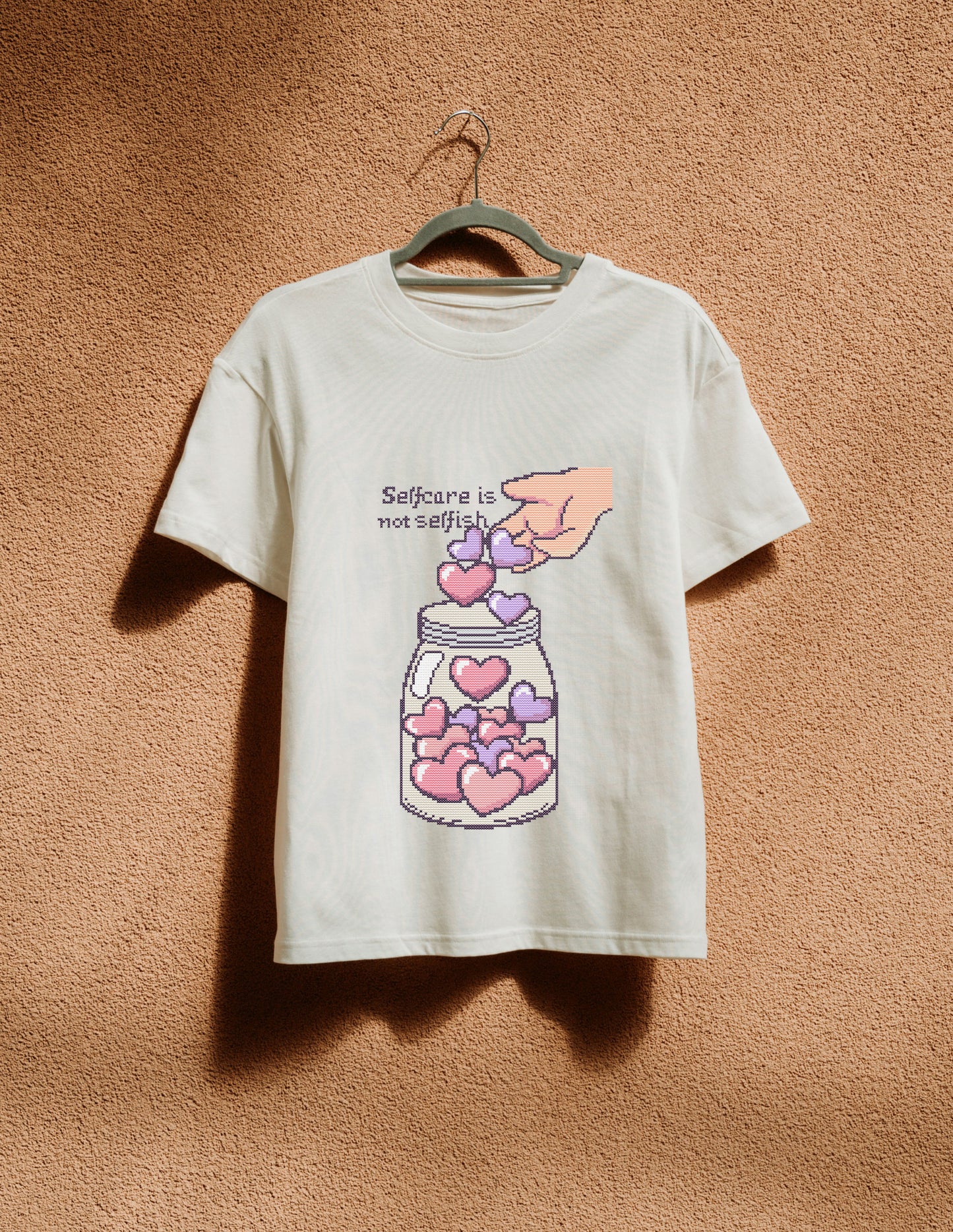 White t-shirt with a graphic design of hearts in a jar and text 'Selfcare is not selfish' on a beige background.
