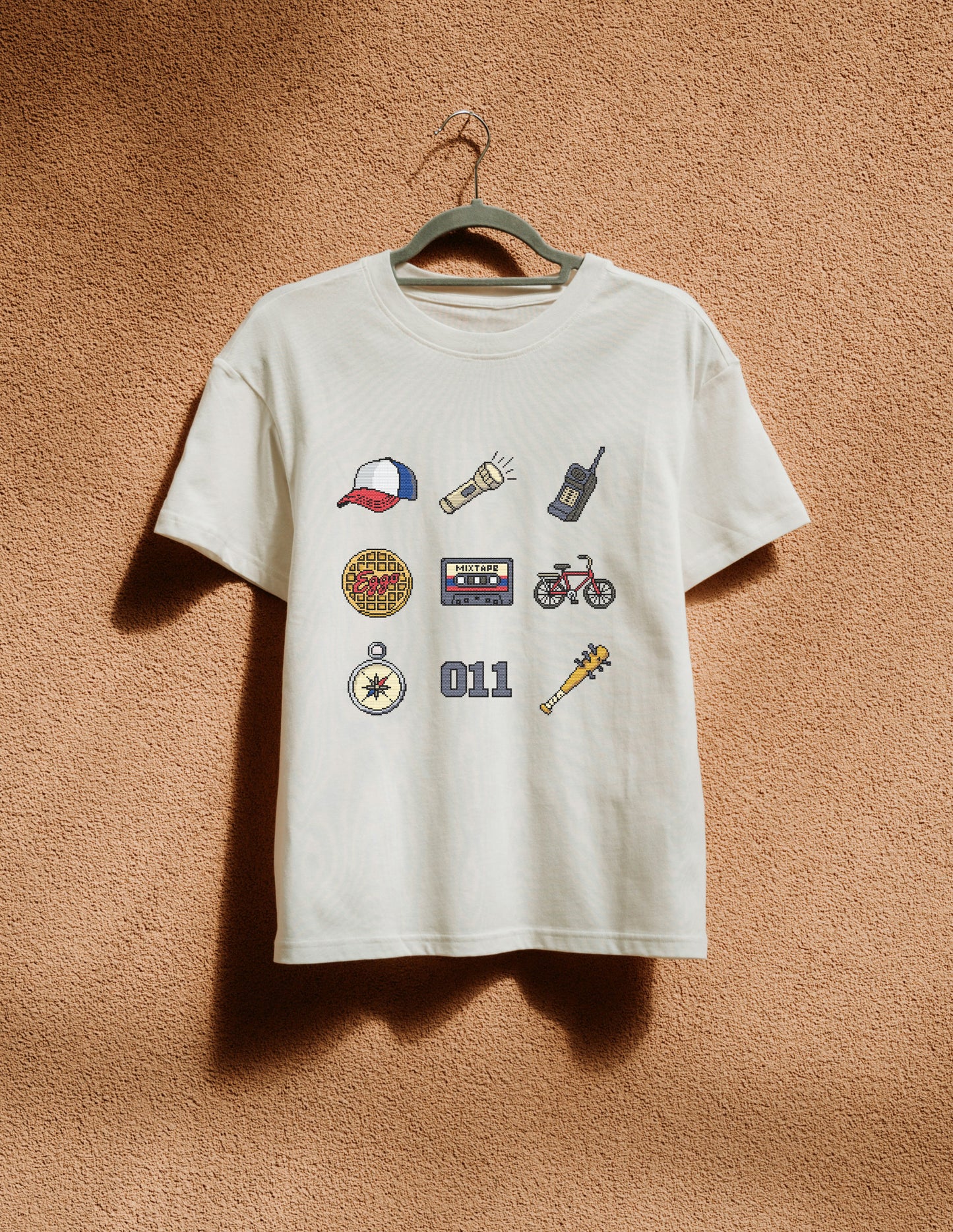 White t-shirt with various graphic designs on a hanger against a brown textured wall.