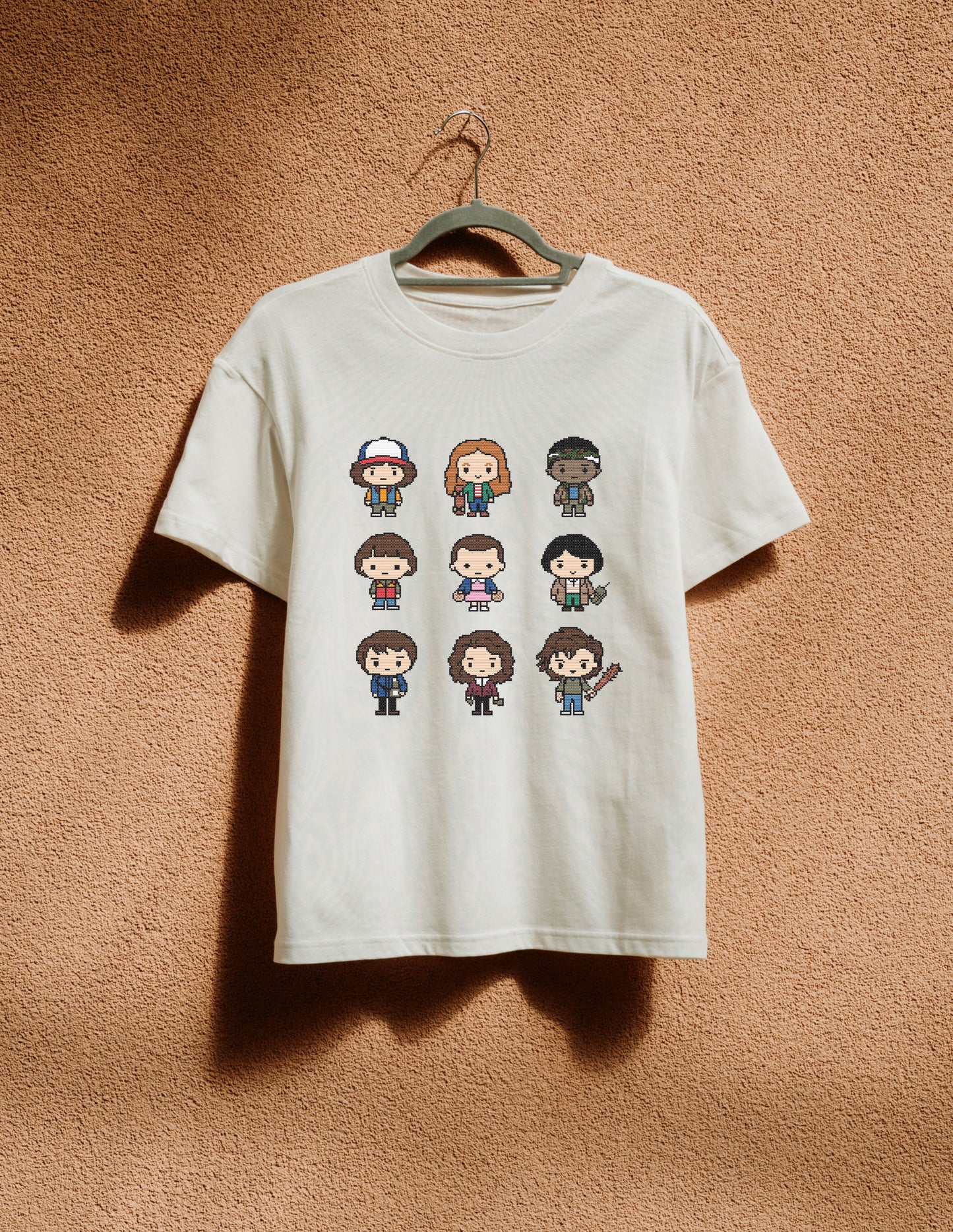 White t-shirt with pixelated character designs on a hanger against a brown textured wall.