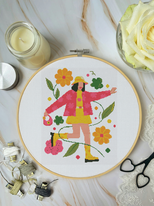 Cross-stitched hoop with a colorful design of a person in a raincoat and wellies, surrounded by crafting materials on a marble surface.