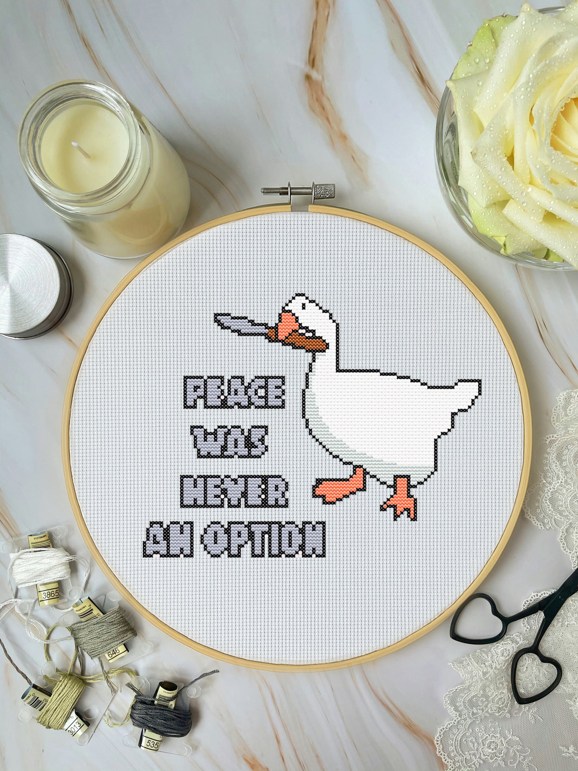 Cross-stitch pattern of a character in an embroidery hoop on a marble surface with a candle and flowers.