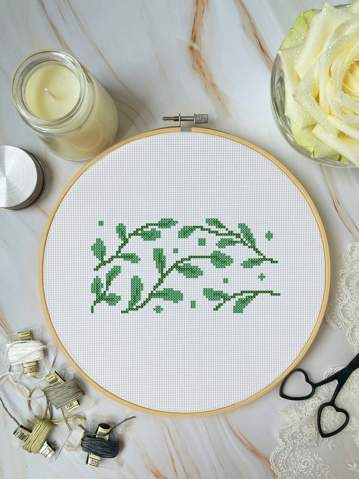 Cross-stitch hoop with green leaf pattern on a marble surface with embroidery supplies.