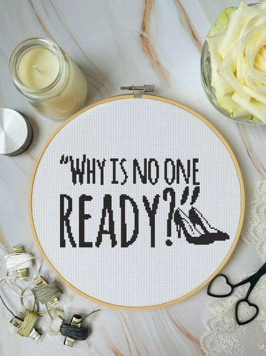Cross-stitched hoop with text 'WHY IS NO ONE READY?' on a marble surface with candles and flowers.
