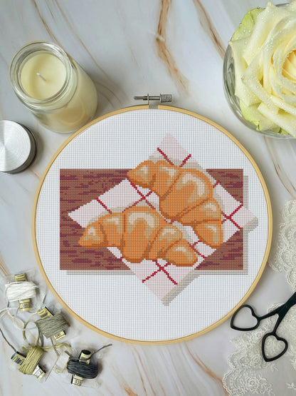 Cross-stitched design of croissants in an embroidery hoop on a marble surface with sewing supplies.