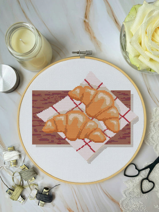 Cross-stitched design of croissants in an embroidery hoop on a marble surface with sewing supplies.