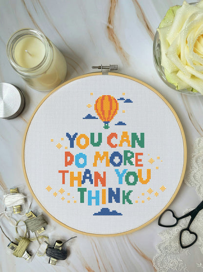 Cross-stitch hoop with colorful motivational quote on a marble surface with crafting supplies.