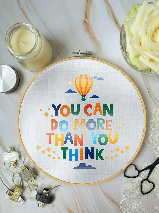 Cross-stitch hoop with colorful motivational quote on a marble surface with crafting supplies.