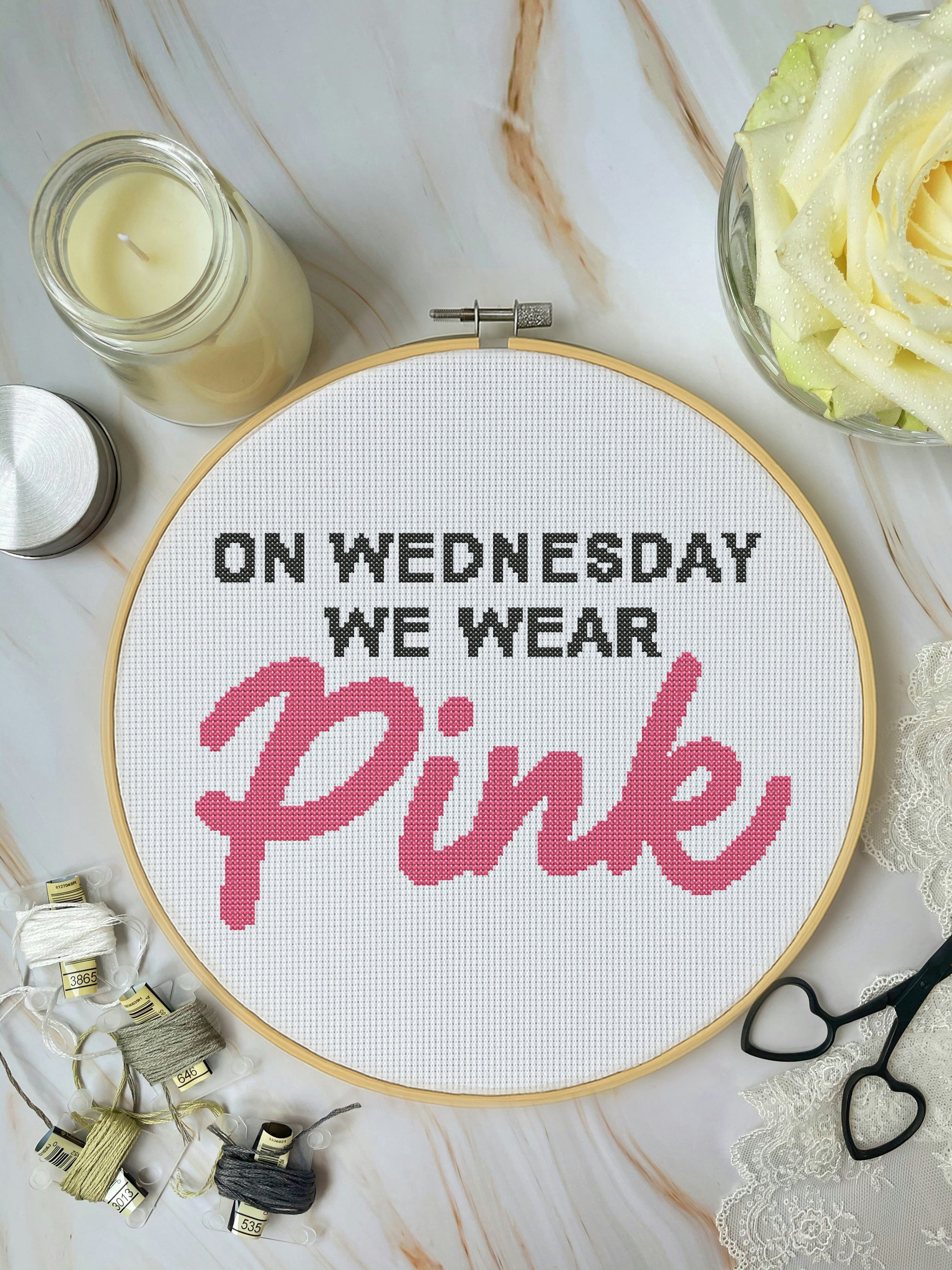 Cross-stitched hoop with 'On Wednesday We Wear Pink' text on a marble surface with candles and flowers.