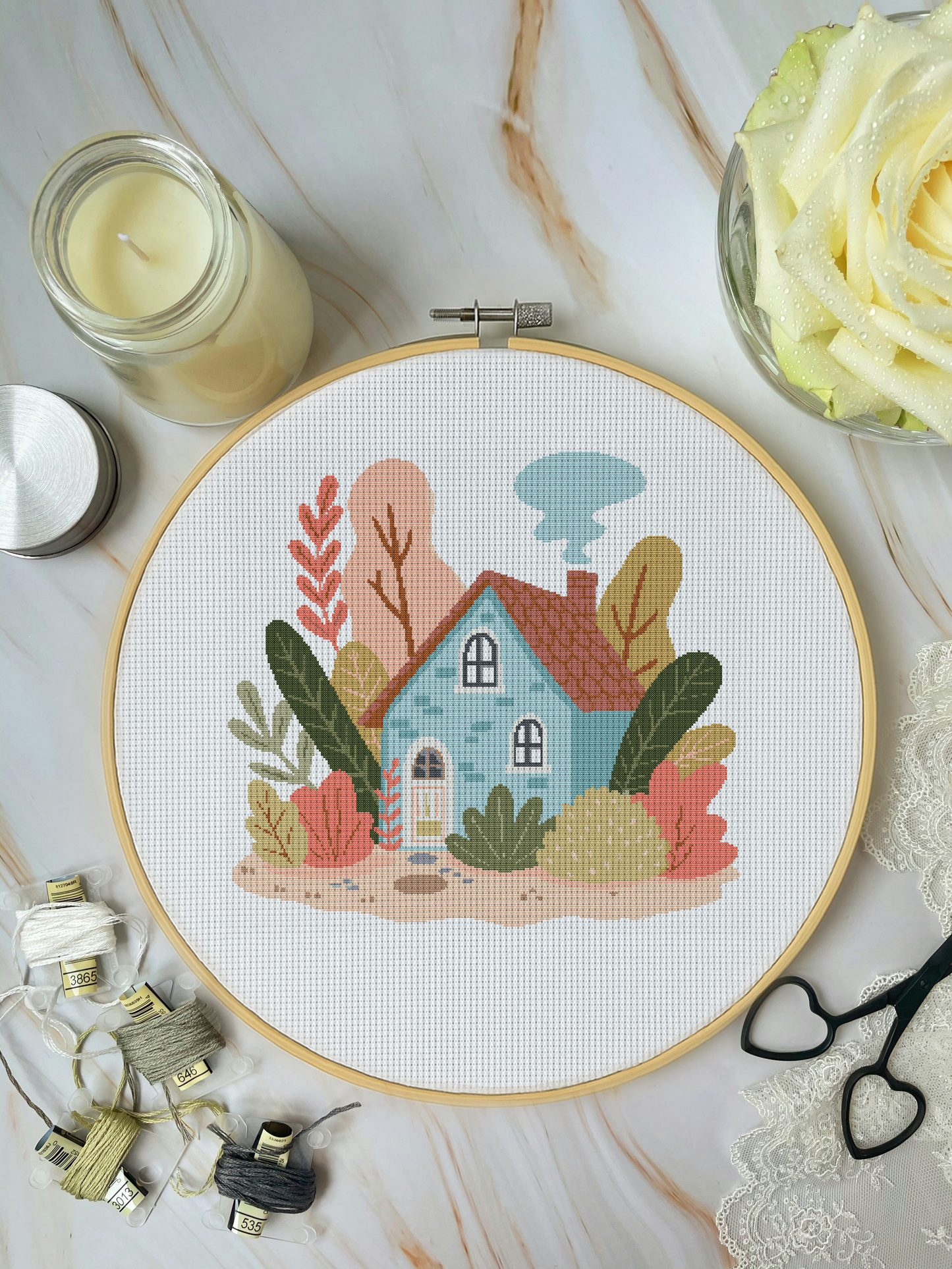 Cross-stitched house design in an embroidery hoop on a marble surface with candles and lace.