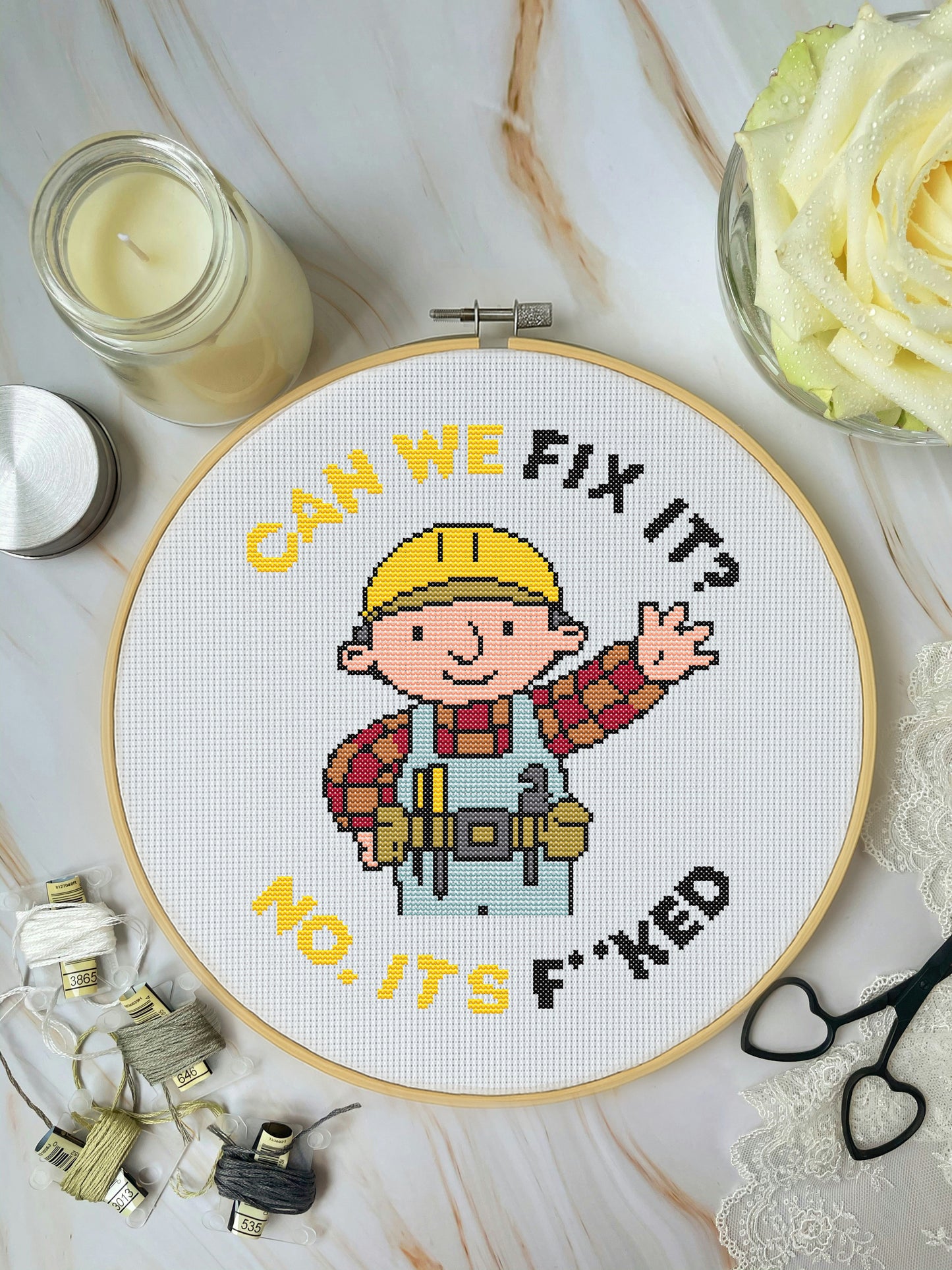 Cross-stitched hoop with cartoon character and text on a marble surface with crafting items.