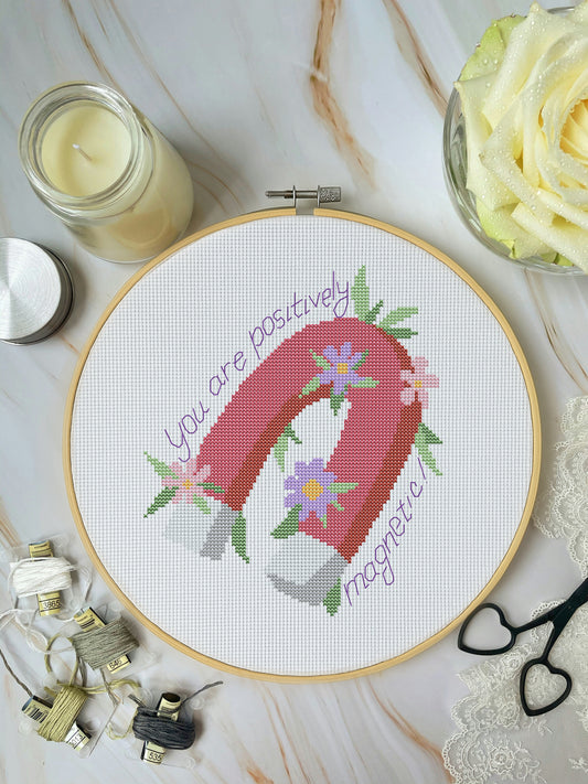 Cross-stitched hoop with 'You are positively magnetic' design on a marble surface with candles and flowers.