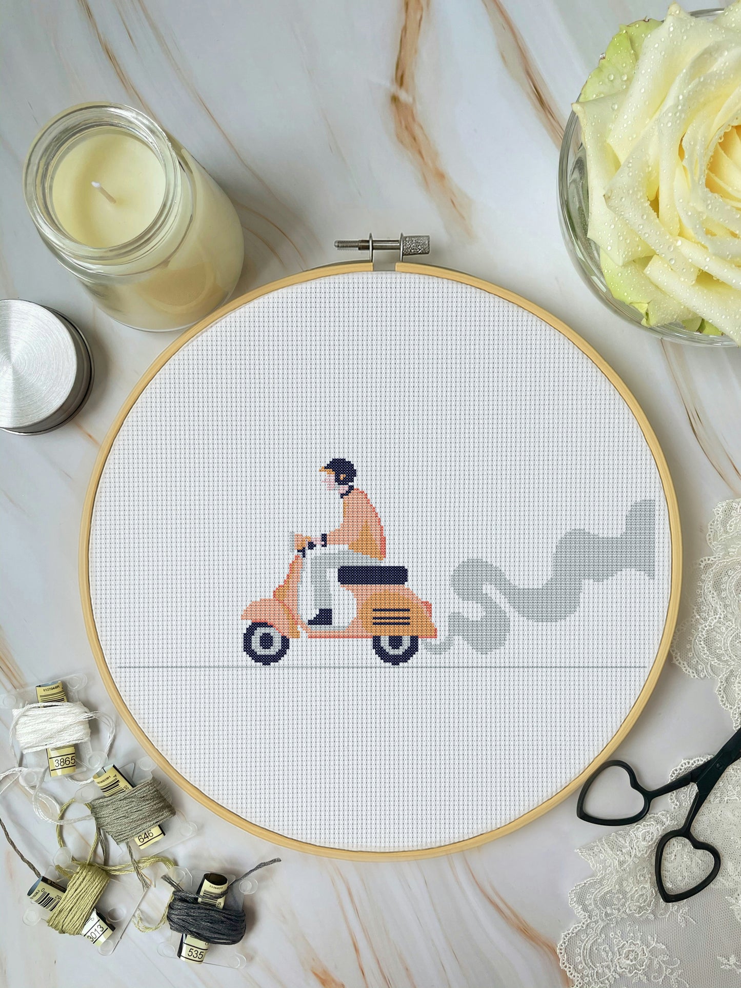 Cross-stitch hoop with a scooter design on a marble surface with crafting materials.