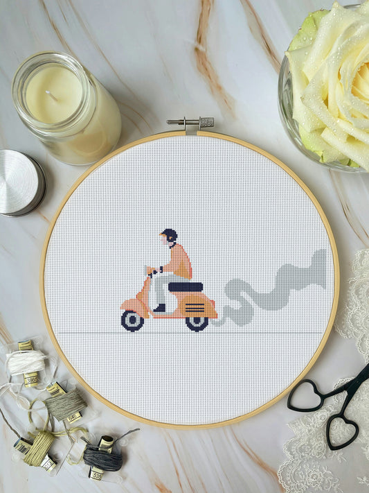 Cross-stitch hoop with a scooter design on a marble surface with crafting materials.