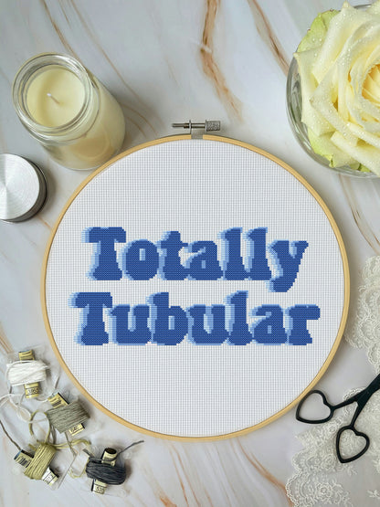 Cross-stitched hoop with 'Totally Tubular' text on a marble surface with crafting supplies.