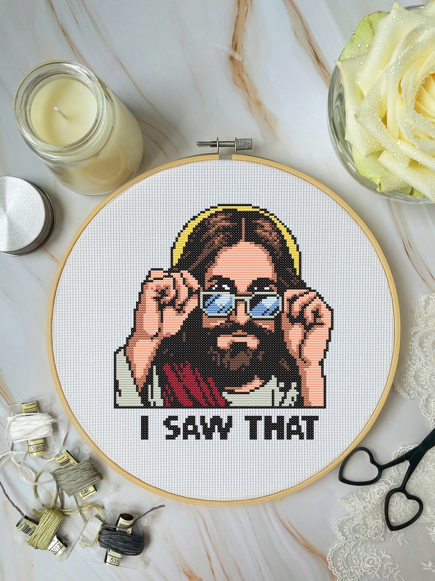 Cross-stitched picture of a person with sunglasses and 'I Saw That' text, surrounded by crafting materials on a marble surface.