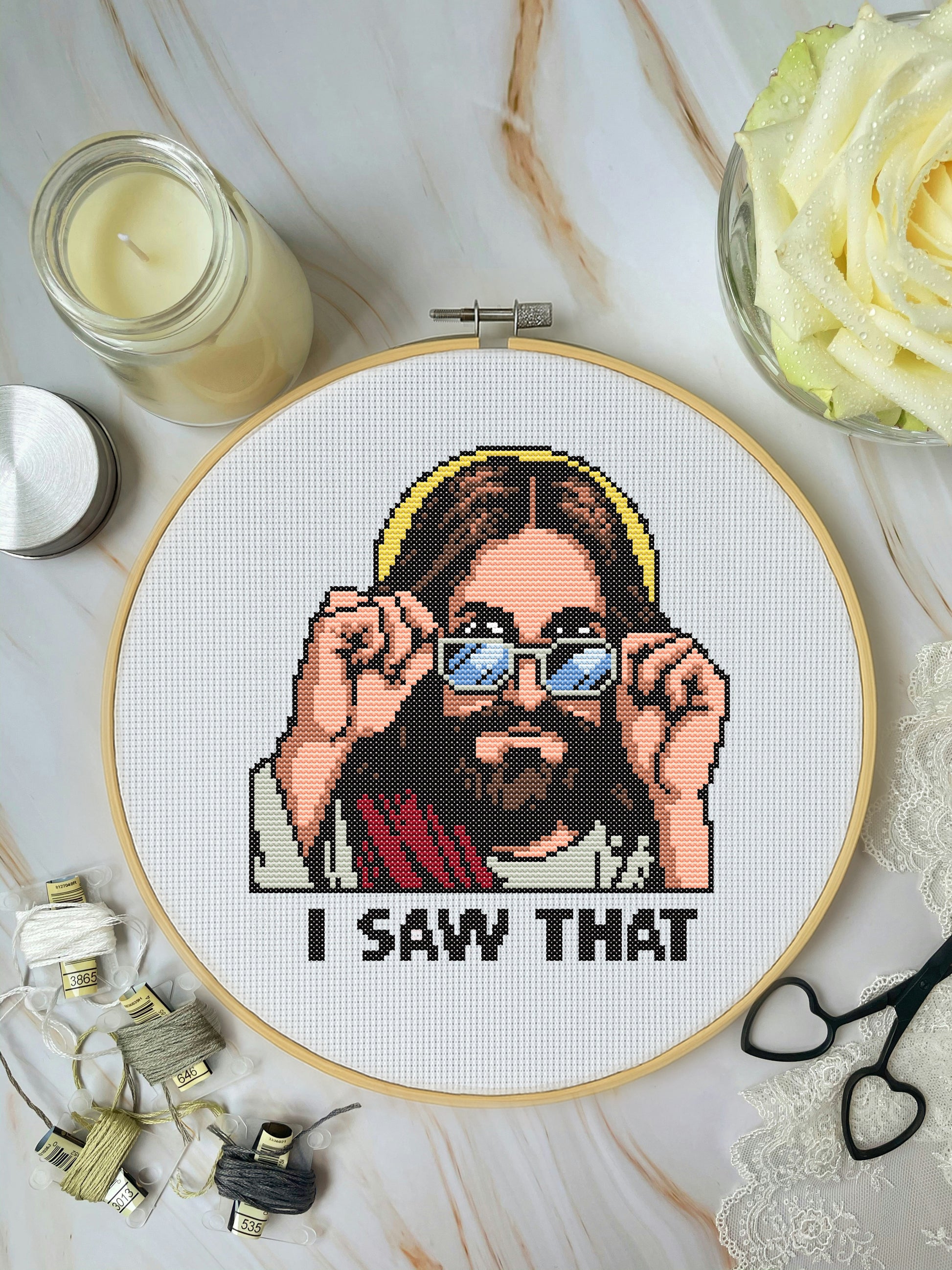 Cross-stitched picture of a person with sunglasses and 'I Saw That' text, surrounded by crafting materials on a marble surface.