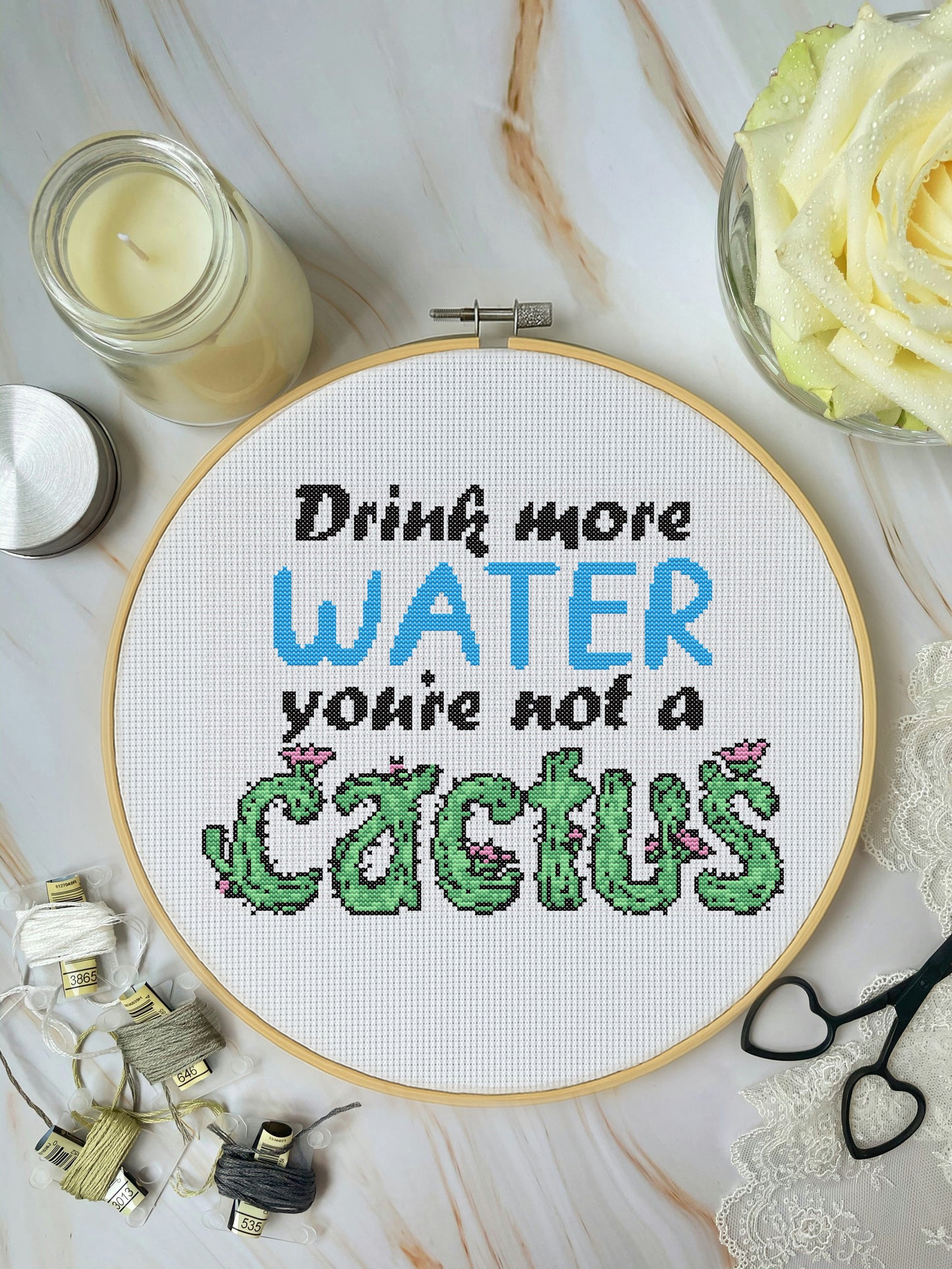 Cross-stitched hoop with 'Drink more water, you're not a cactus' text on a marble surface with candles and flowers.