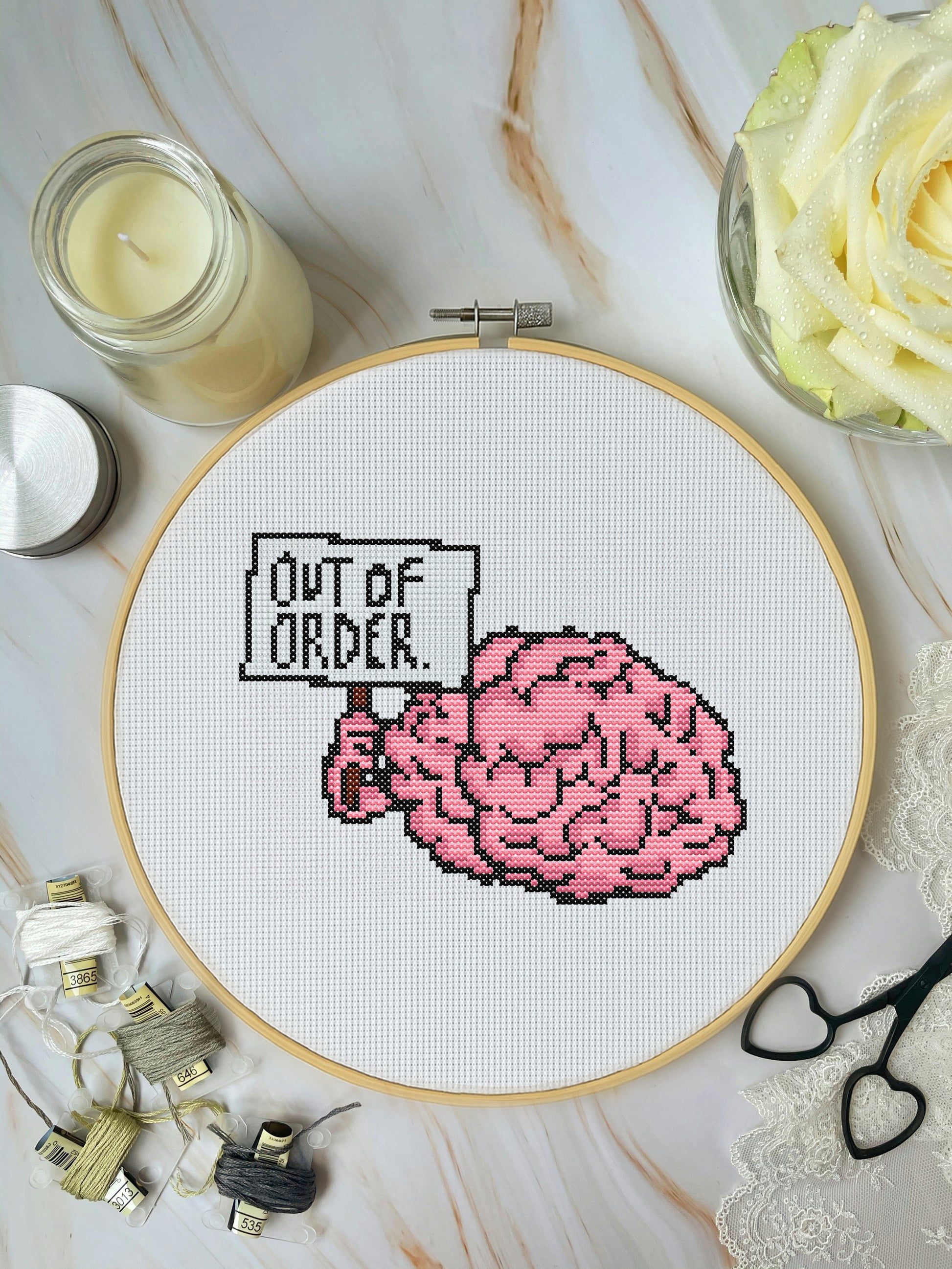 Cross-stitched design of a brain with 'Out of Order' text on a marble surface with crafting materials.