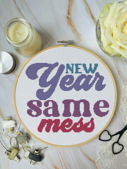 Cross-stitched hoop with 'New Year Same Mess' text on a marble surface with candles and flowers.