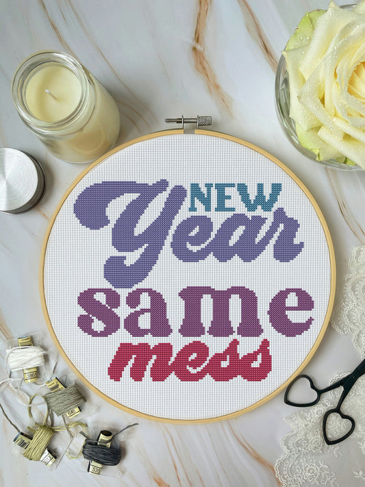Cross-stitched hoop with 'New Year Same Mess' text on a marble surface with candles and flowers.