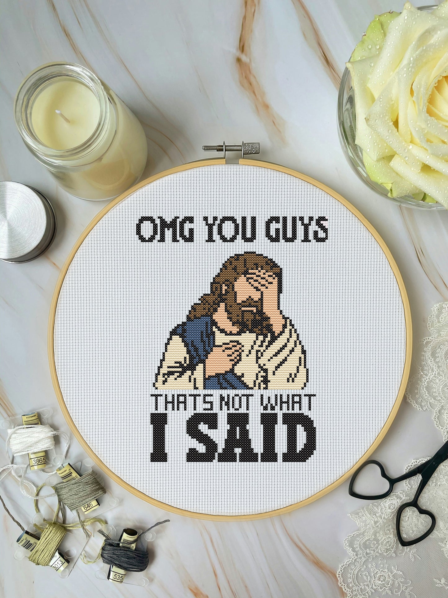 Cross-stitched hoop with humorous design of a character and text, surrounded by crafting supplies on a marble surface.