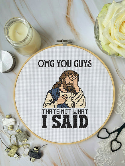 Cross-stitched hoop with humorous design of a character and text, surrounded by crafting supplies on a marble surface.