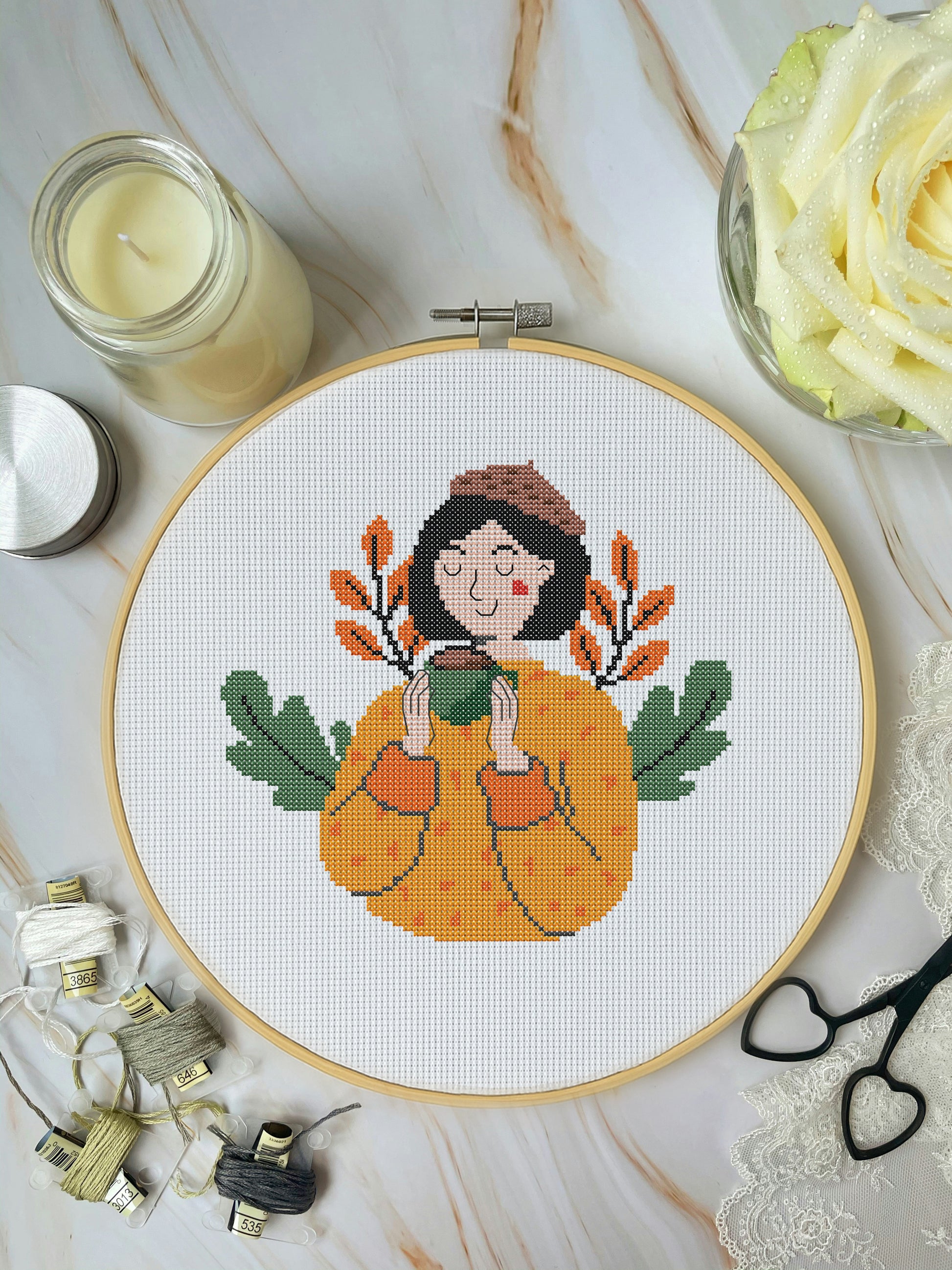 Cross-stitched design of a person in a pumpkin with autumn leaves on a marble surface.