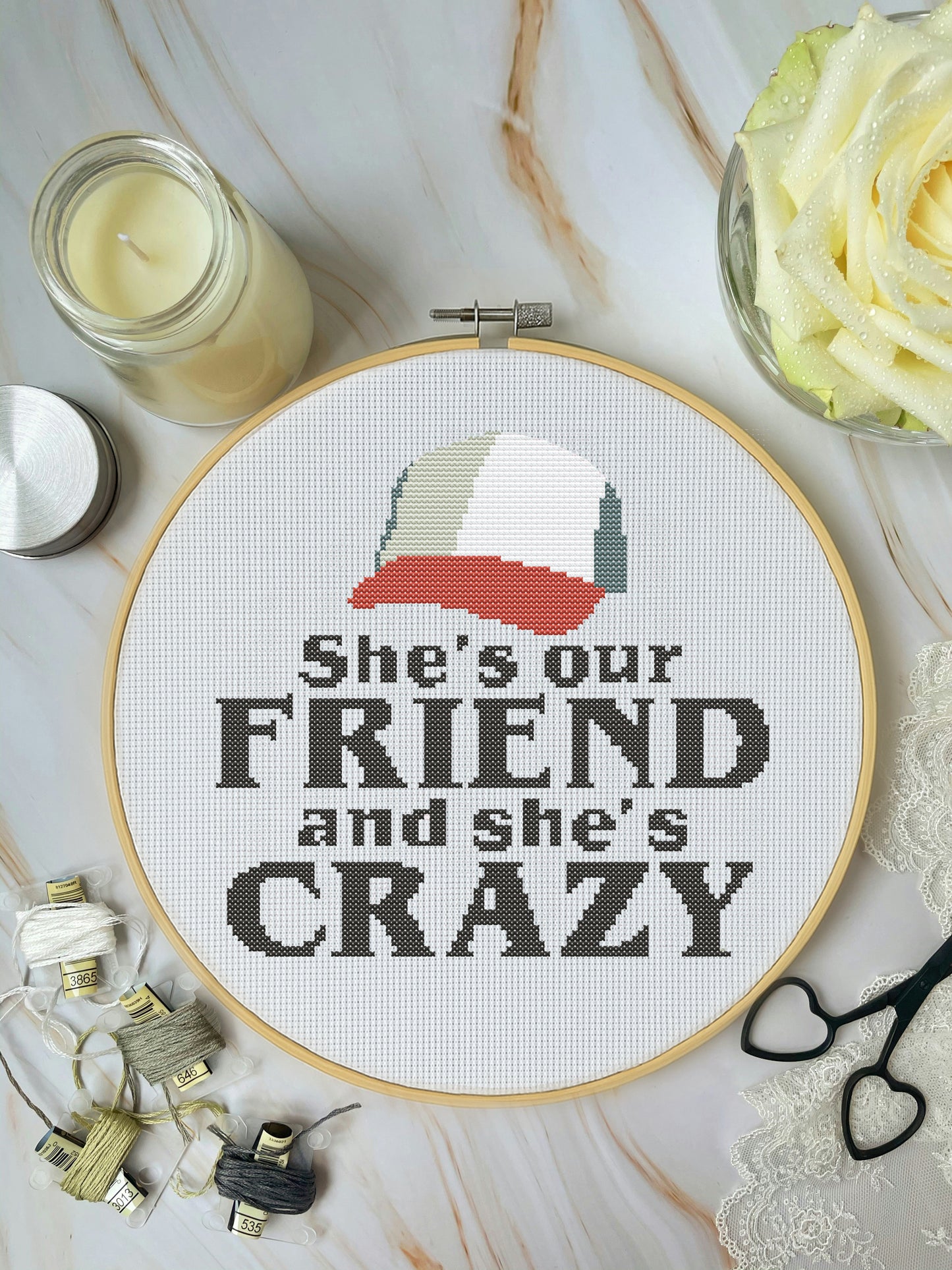 Cross-stitched hoop with text 'She's our FRIEND and she's CRAZY' on a marble surface with decorative items.
