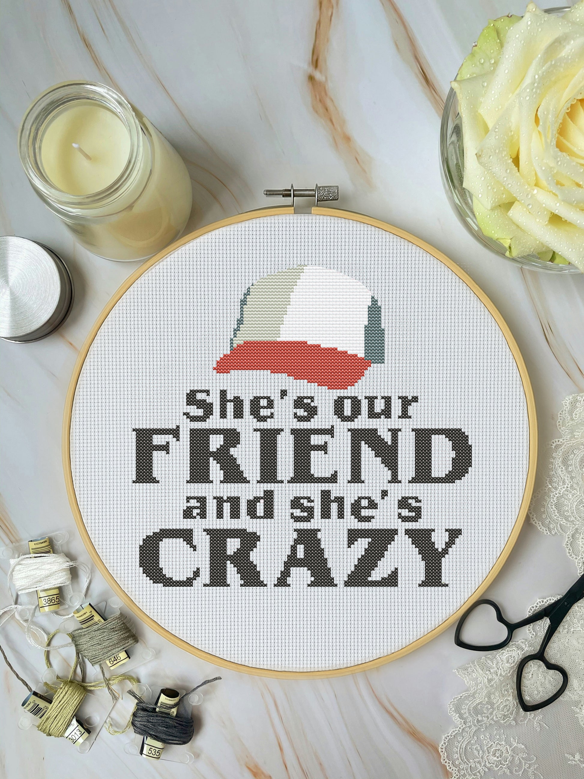 Cross-stitched hoop with text 'She's our FRIEND and she's CRAZY' on a marble surface with decorative items.