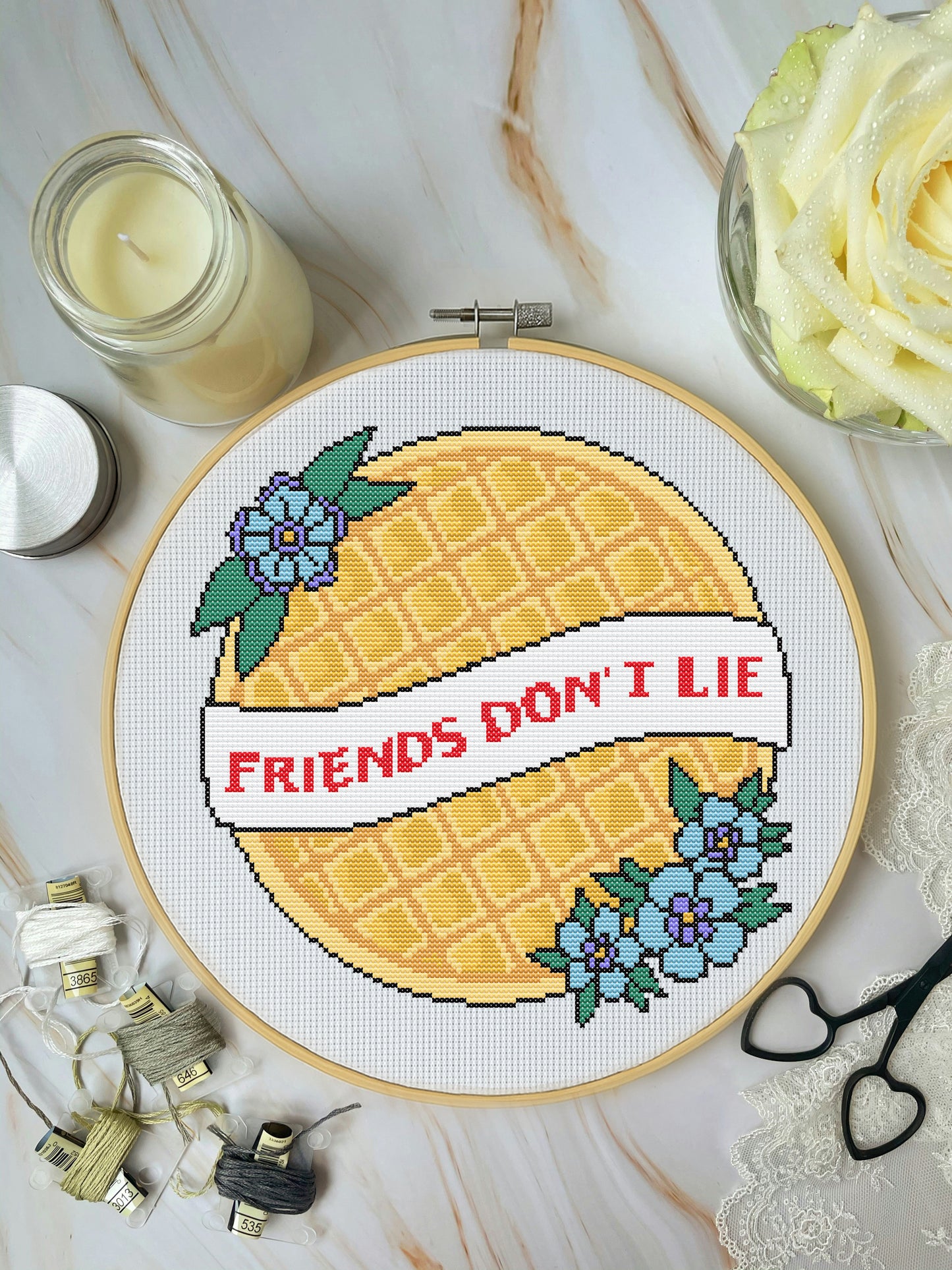 Cross-stitched pineapple with 'Friends Don't Lie' text on a wooden hoop, surrounded by crafting materials.
