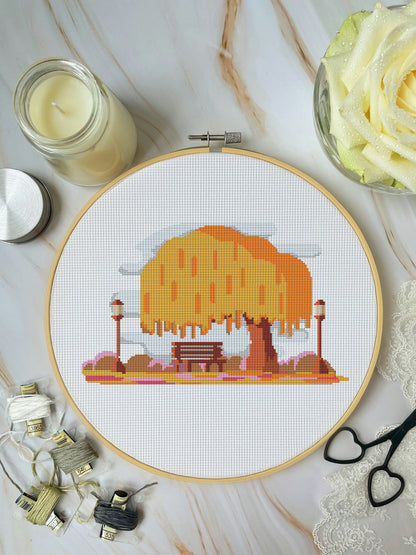Cross-stitch hoop with a completed design of a tree and bench on a marble surface with sewing supplies.