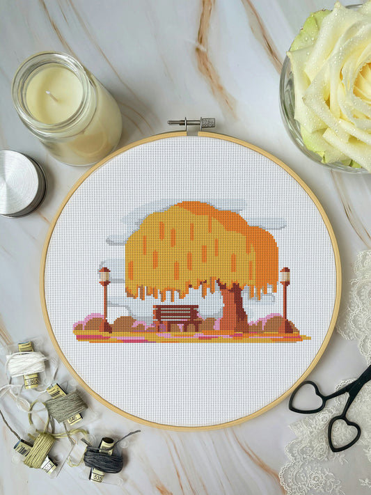 Cross-stitch hoop with a completed design of a tree and bench on a marble surface with sewing supplies.