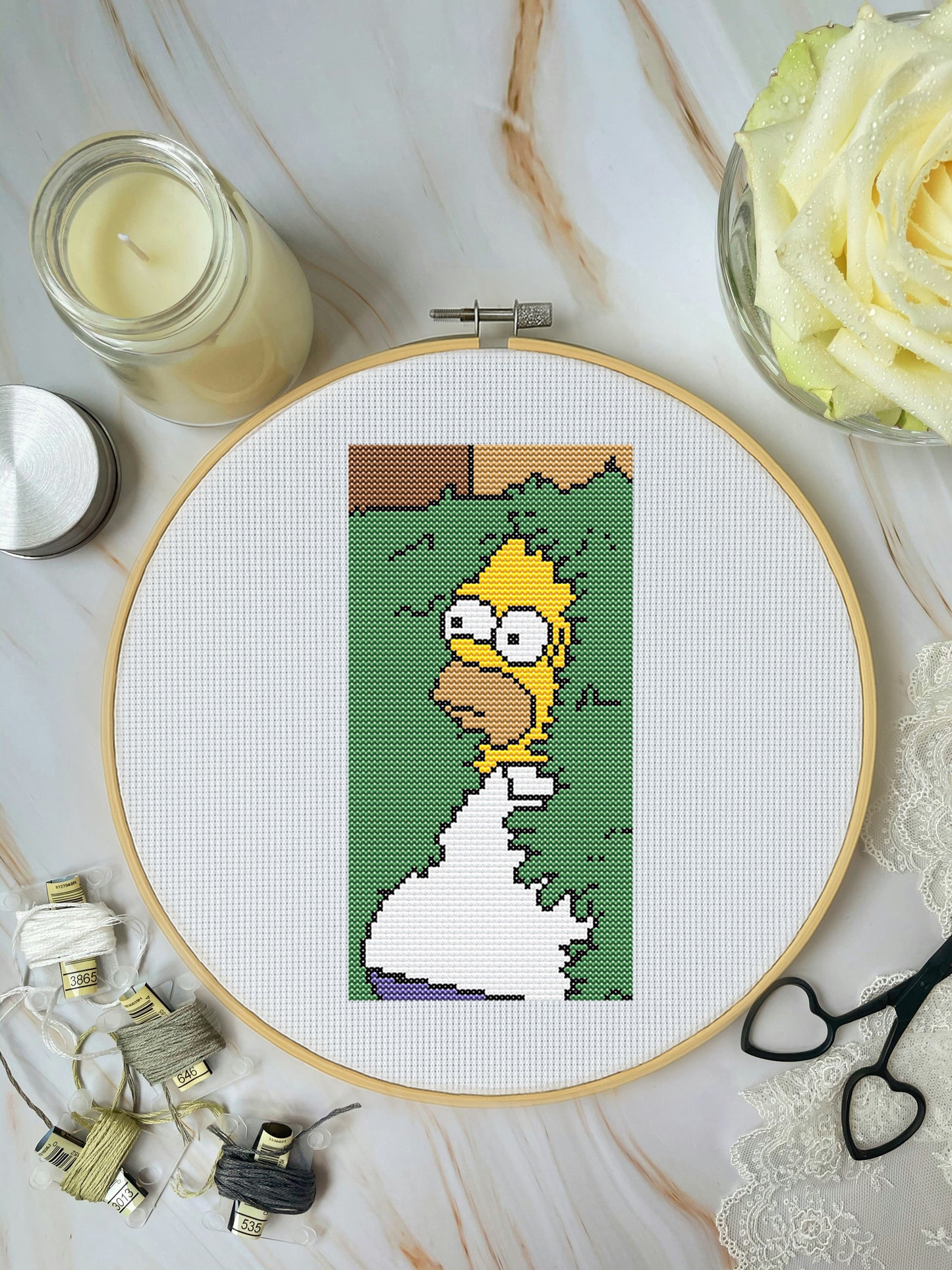 Cross-stitch pattern of a character in an embroidery hoop on a marble surface with a candle and flowers.