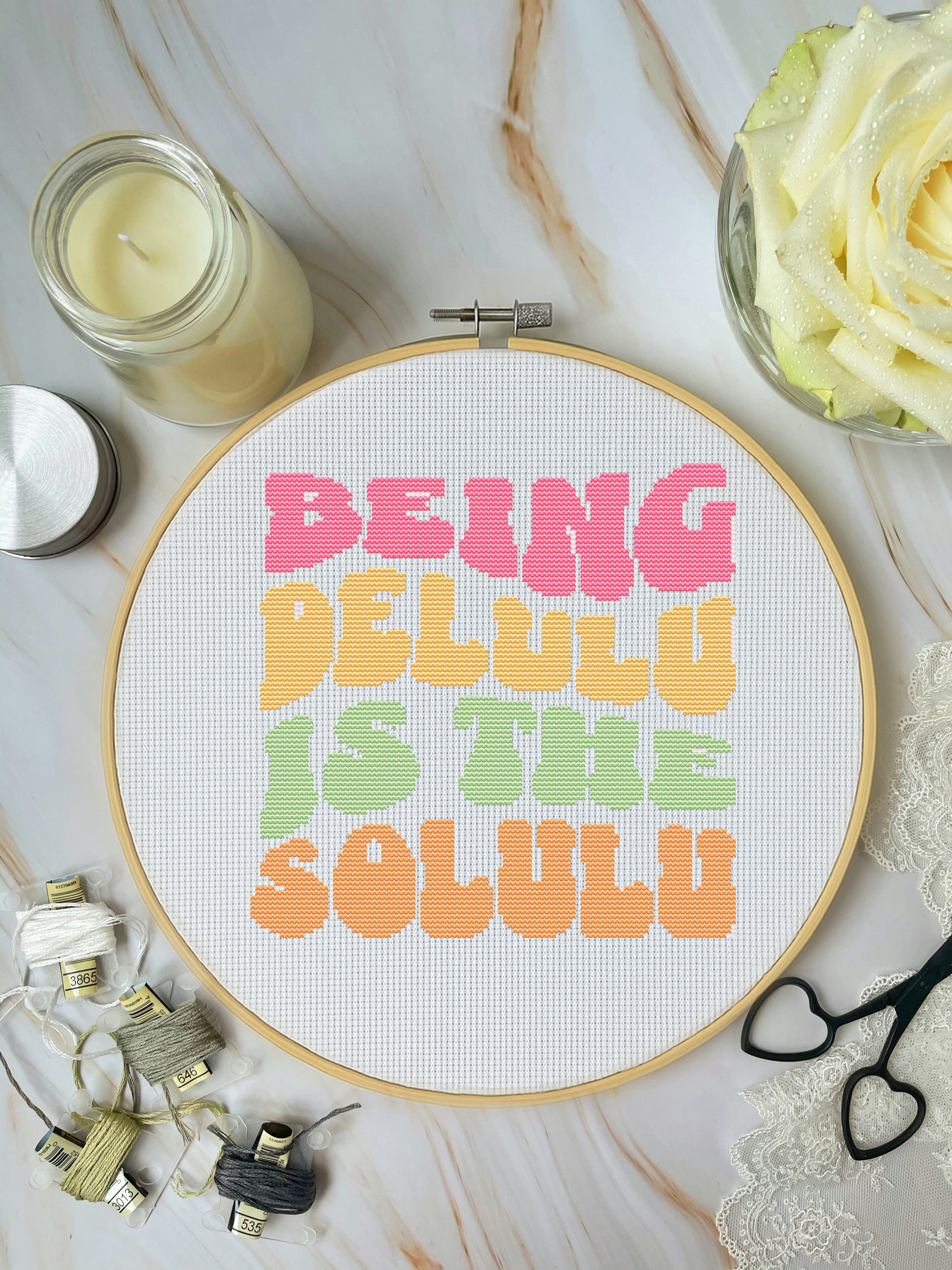 Cross-stitch hoop with colorful text on a marble surface with crafting supplies.