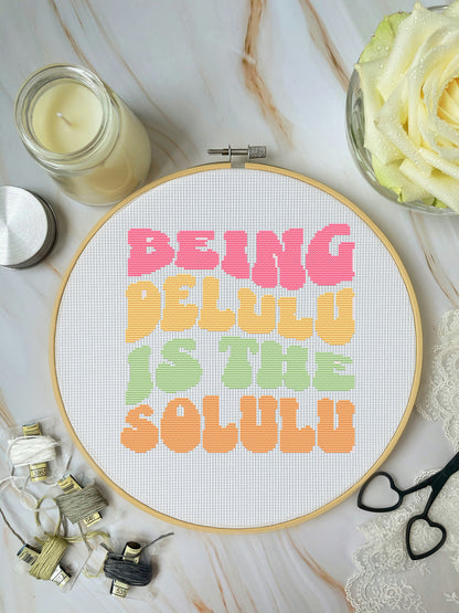 Cross-stitch hoop with colorful text on a marble surface with crafting supplies.