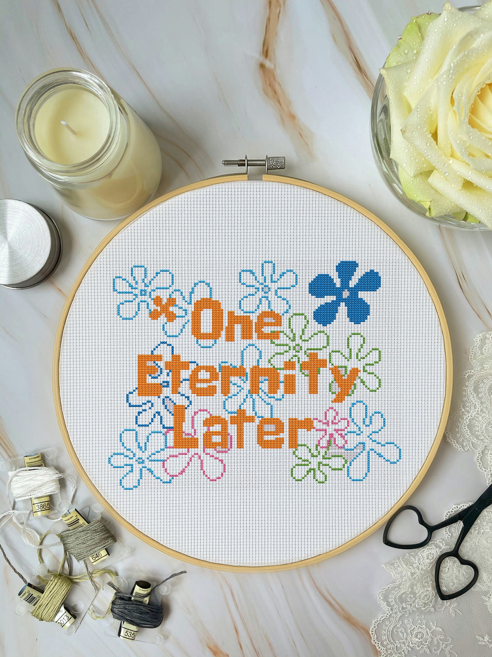 Cross-stitched hoop with 'One Eternity Later' text and flowers on a marble surface with candles and lace.