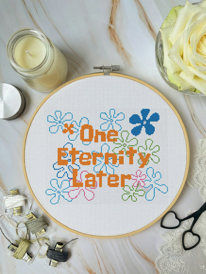 Cross-stitched hoop with 'One Eternity Later' text and flowers on a marble surface with candles and lace.