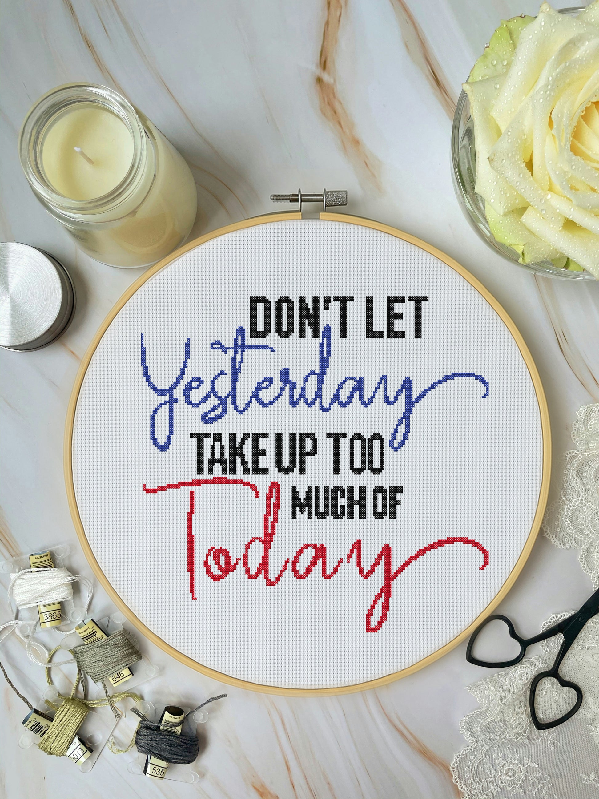 Cross-stitched hoop with motivational quote on a marble surface with candles and flowers.