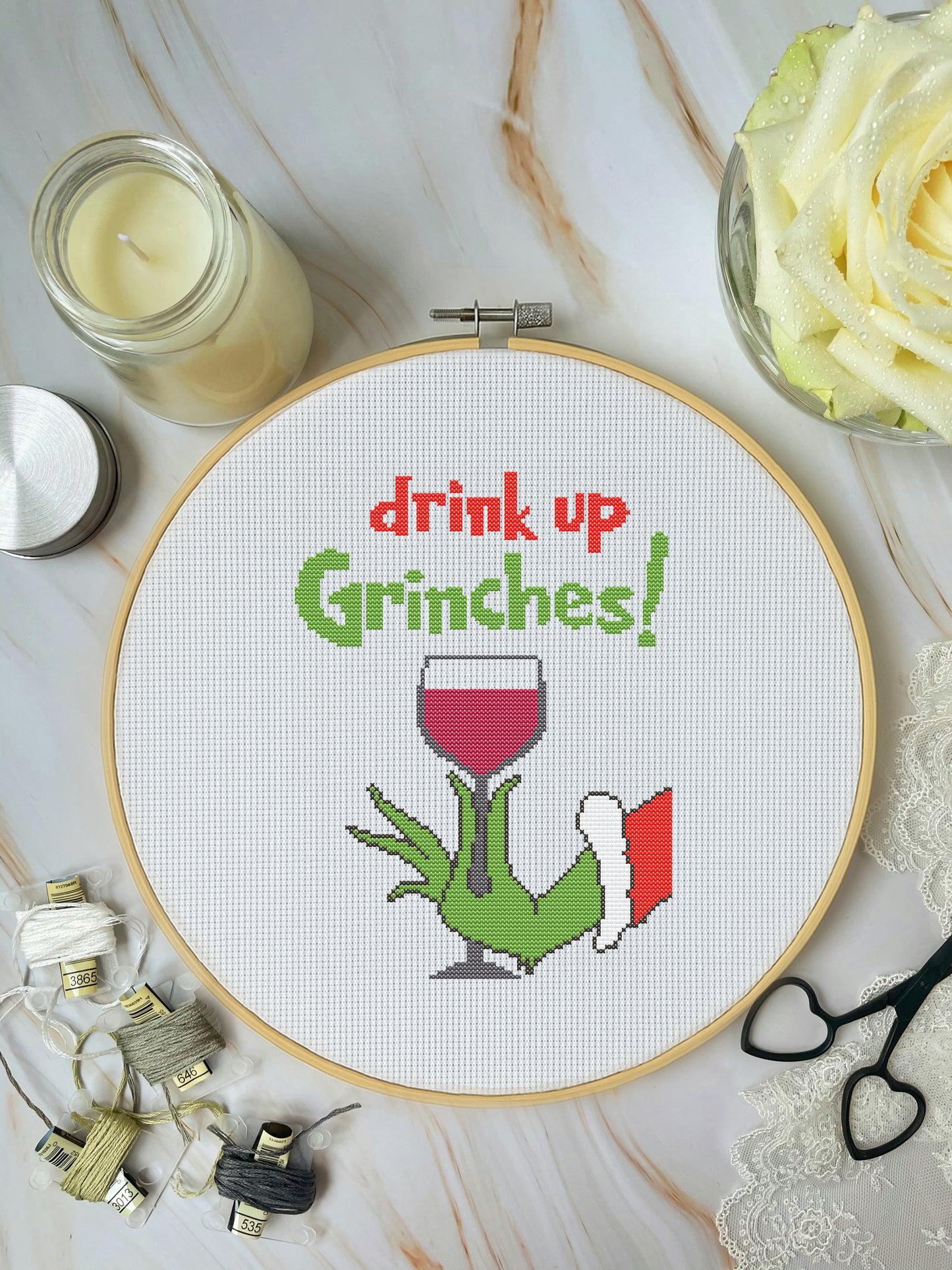 Cross-stitched design with 'drink up Grinches!' text and a Grinch holding a wine glass, surrounded by crafting materials on a marble surface.