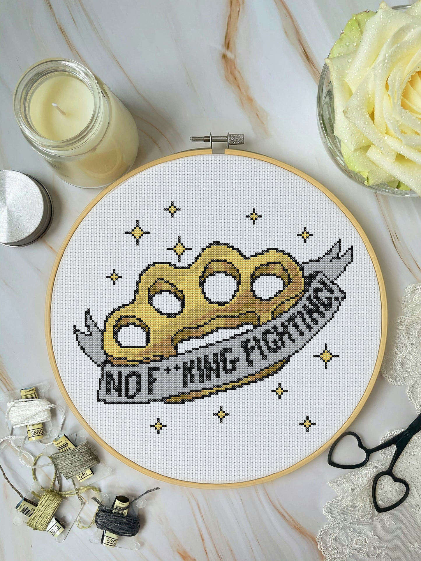 Cross-stitched design of a knuckle duster with 'No F*cking Fighting!' text in a hoop, surrounded by crafting materials on a marble surface.