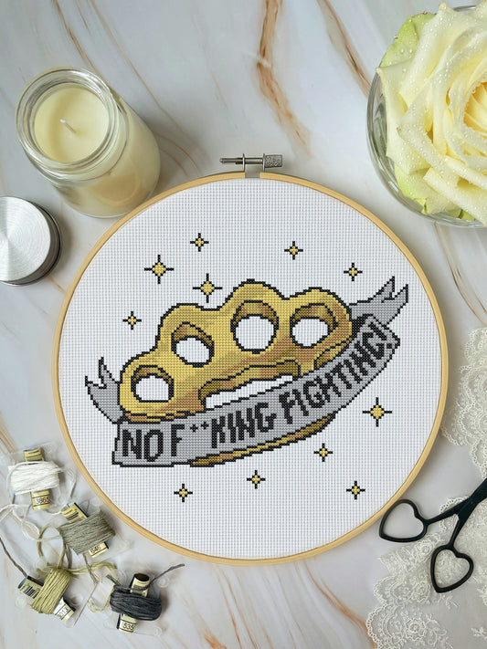 Cross-stitched design of a knuckle duster with 'No F*cking Fighting!' text in a hoop, surrounded by crafting materials on a marble surface.