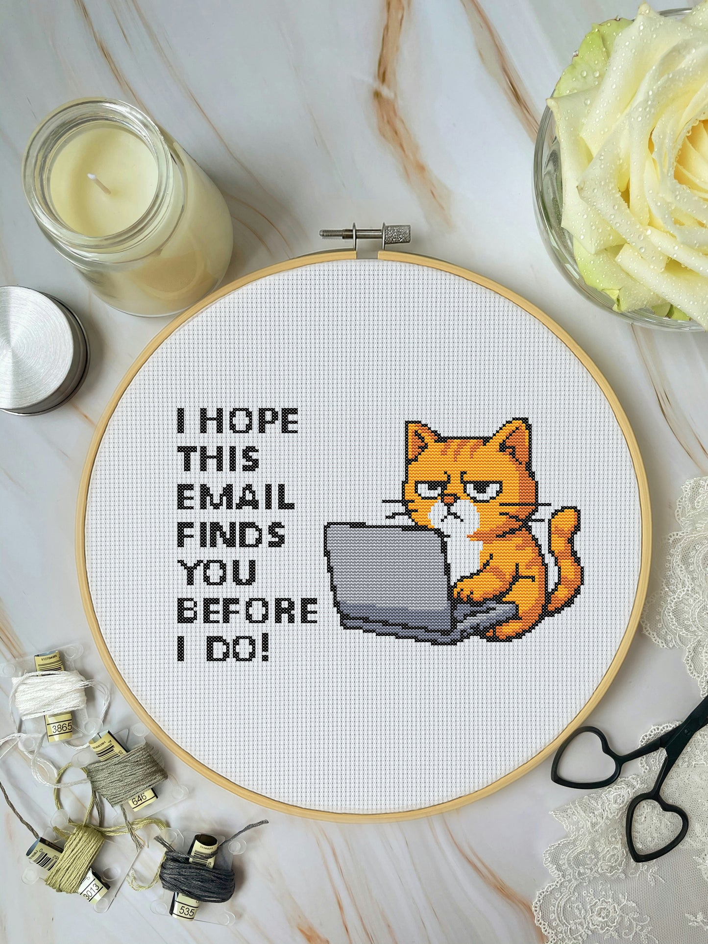 Embroidery hoop with a cat on a laptop and text, surrounded by crafting supplies on a marble surface.