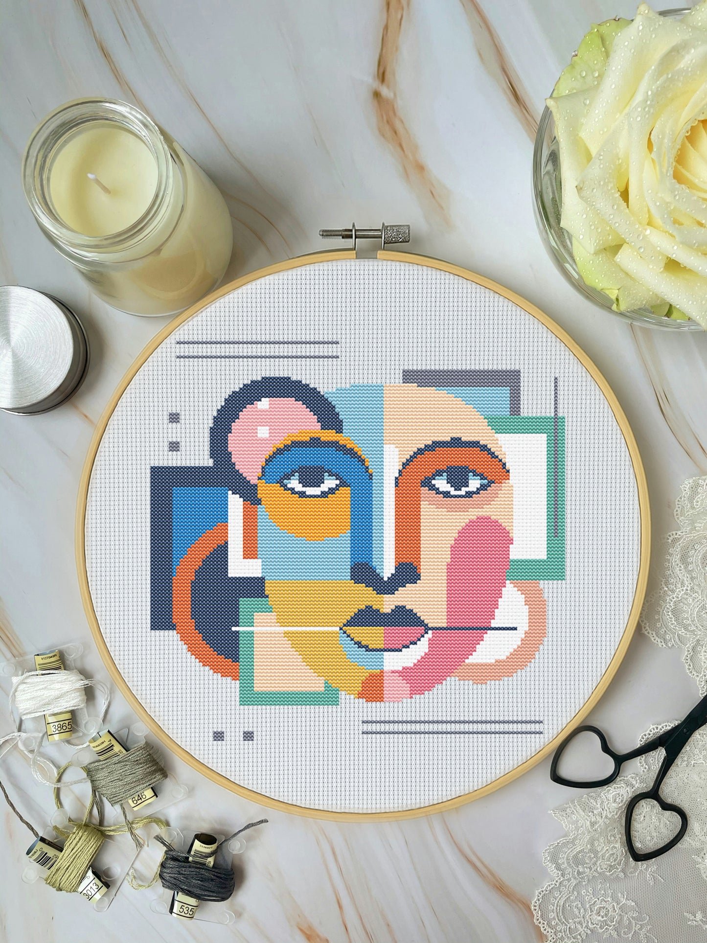 Cross-stitching hoop with geometric face design on a marble surface with crafting materials.