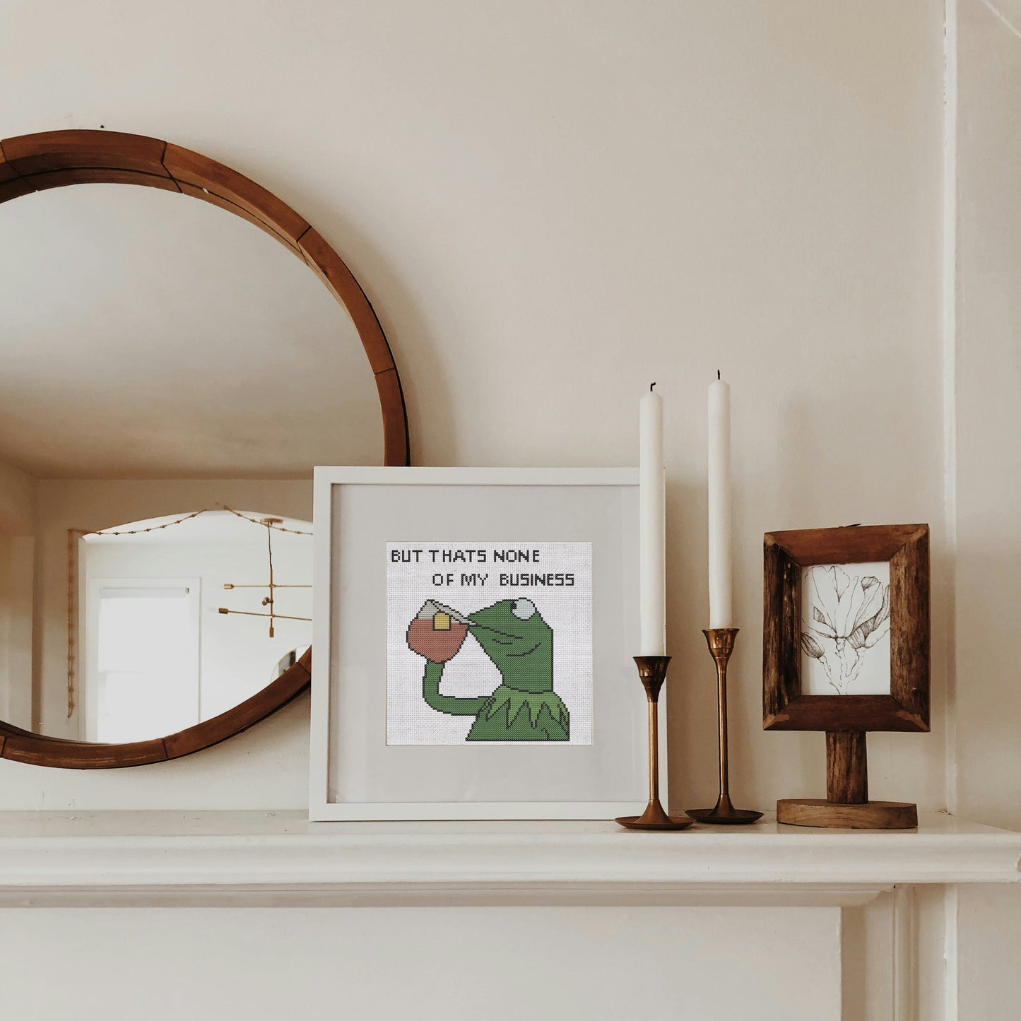 Framed artwork with a green character and text on a shelf with candles and a mirror.