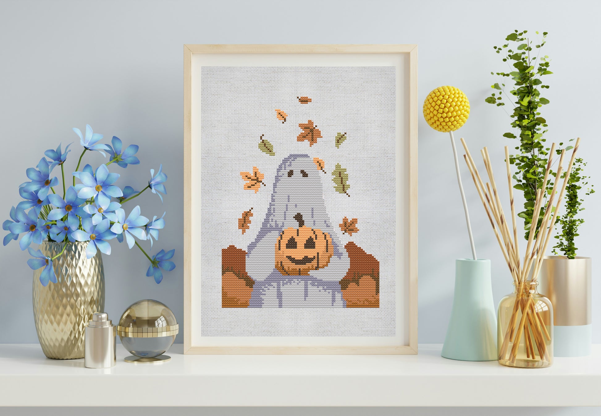 Framed Halloween-themed cross-stitch art of a ghost with a pumpkin on a shelf with decorative items.