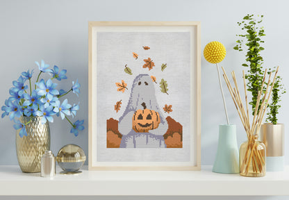 Framed Halloween-themed cross-stitch art of a ghost with a pumpkin on a shelf with decorative items.