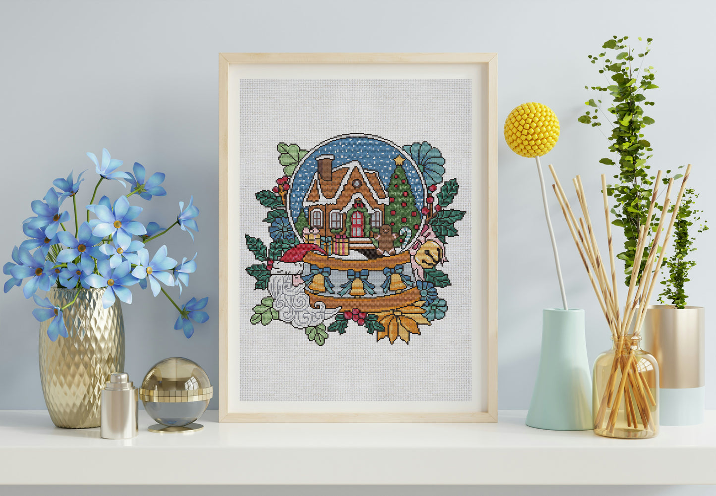 Framed cross-stitch art of a snowy landscape with a house on a shelf with decorative items.