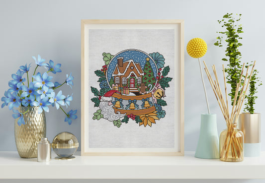 Framed cross-stitch art of a snowy landscape with a house on a shelf with decorative items.