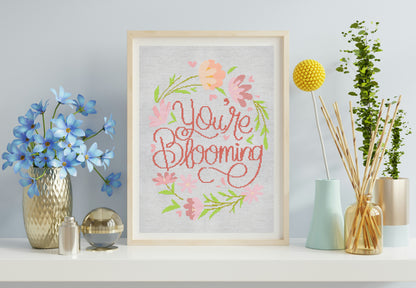 Framed artwork with 'You're Blooming' text and floral design on a shelf with decorative items.