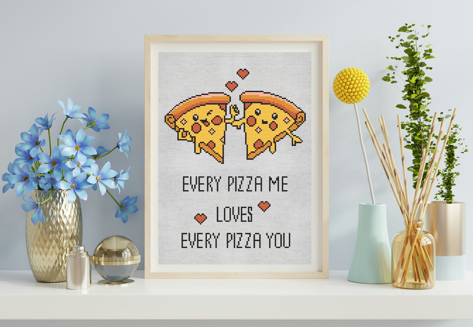 Cross-stitched pizza design with text in a wooden frame on a shelf with decorative items.